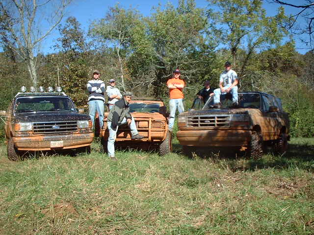 Three muddy Ford trucks and SUVs with men posing in a grassy field
