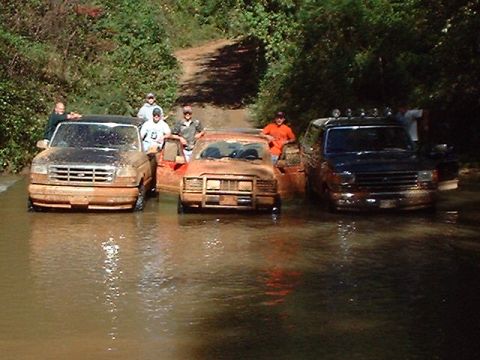 1990 Ford Bronco - Three SUVs, including a 1990 Ford Bronco, navigate a muddy water crossing on a dirt road.