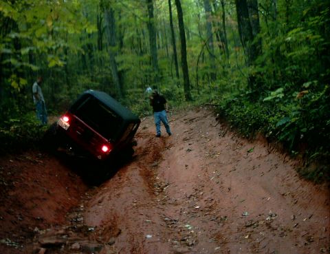 1990 Ford Bronco - A red 1990 Ford Bronco navigates a challenging muddy incline in a wooded area.