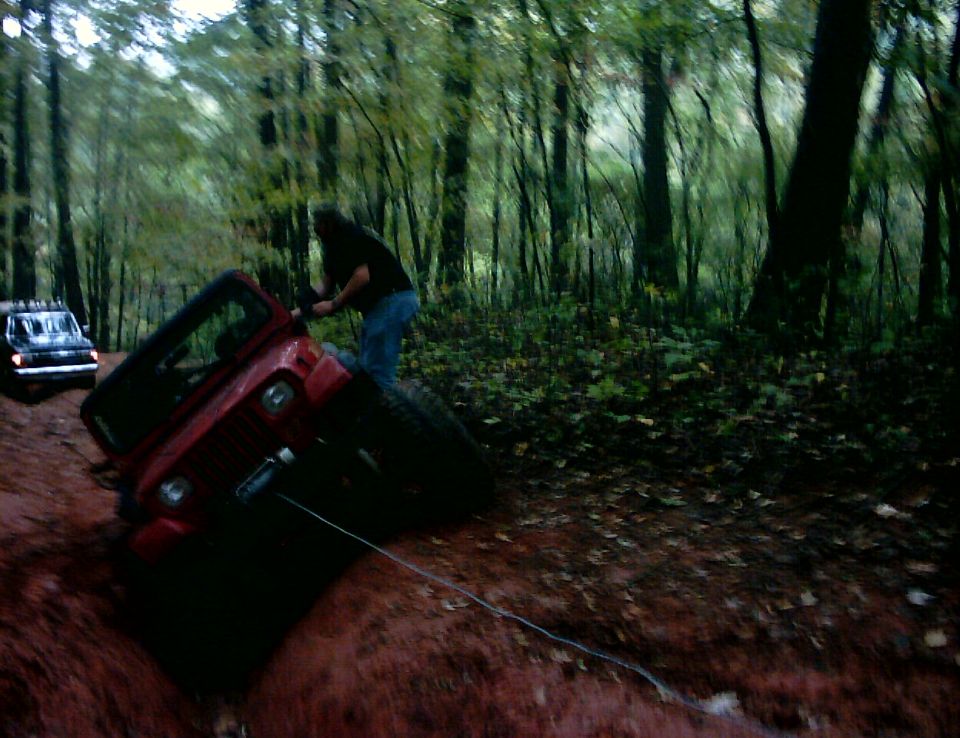 Red Jeep tilted on dirt embankment with person on front tire in forest