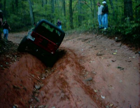 1990 Ford Bronco - A Jeep Wrangler navigates a steep, muddy incline in a wooded area.