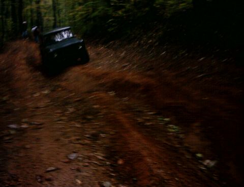 1990 Ford Bronco - A dark SUV navigates a muddy forest road, its tires kicking up dirt.
