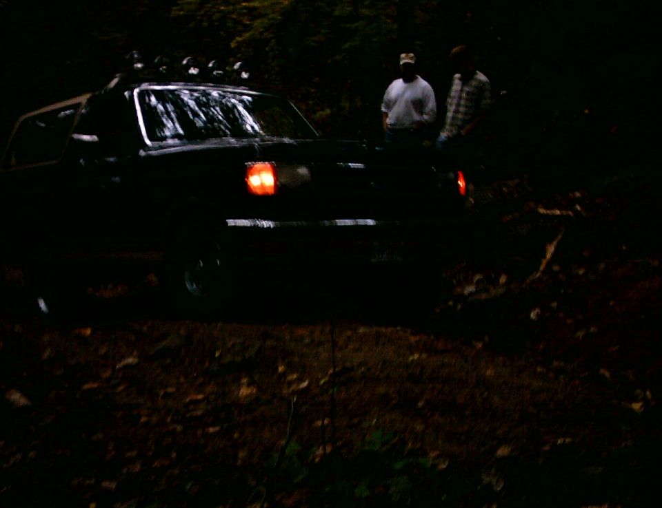 Dark Ford Bronco with roof light bar and glowing tail lights on a muddy trail at dusk