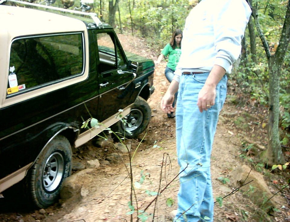 Black and tan Ford Bronco on a rocky wooded trail, rear three-quarter view
