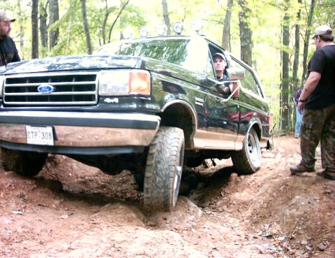 1990 Ford Bronco - A black Ford Bronco navigates a challenging off-road trail, its front tire elevated on a dirt mound.