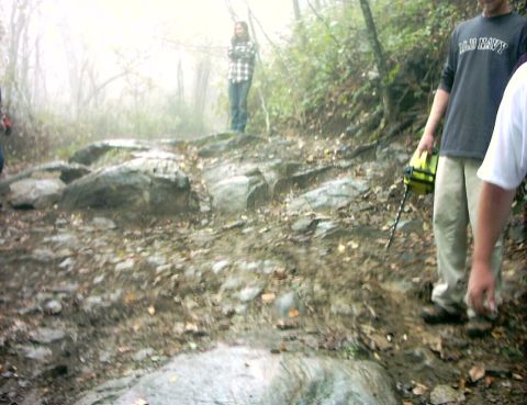 1990 Ford Bronco - A group of people navigate a challenging, muddy, and rocky trail in a foggy, wooded environment.