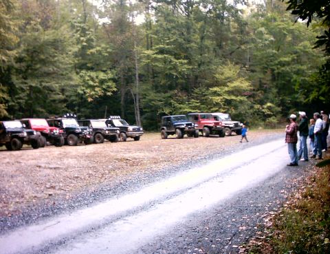 1990 Ford Bronco - A group of off-road vehicles are gathered in a wooded area, with people observing from the roadside.