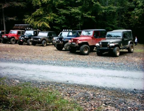 1990 Ford Bronco - Several off-road vehicles are parked in a line on a gravel lot, with a wooded area behind them.
