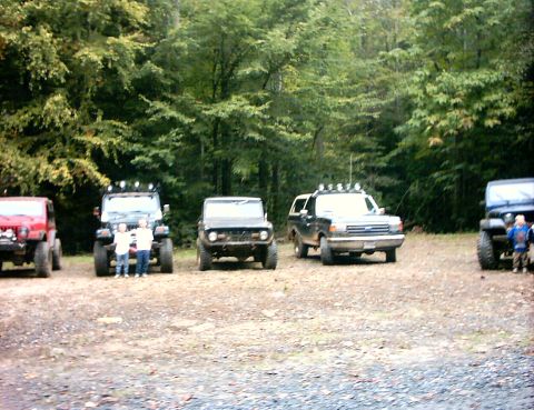 1990 Ford Bronco - Several off-road vehicles are parked in a dirt lot with trees in the background. One vehicle displays a "SUPER MOTORS" watermark.