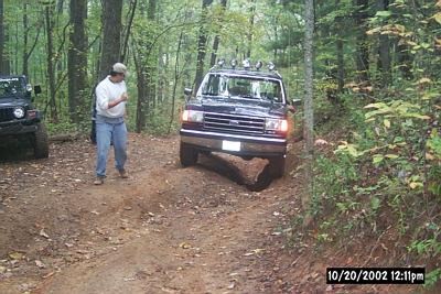 1990 Ford Bronco - A dark Ford F-Series truck navigates a wooded trail, with another vehicle visible to the left.