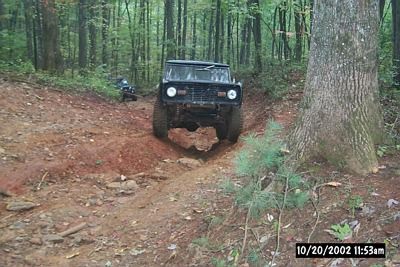 1990 Ford Bronco - An off-road vehicle navigates a steep, rutted dirt path surrounded by trees.