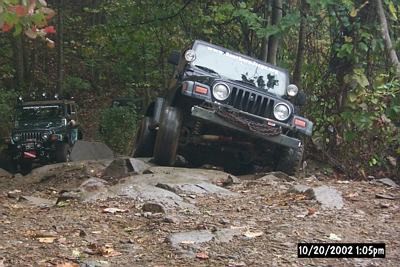 1990 Ford Bronco - Two Jeep Wranglers navigate a challenging off-road trail with rocky obstacles and dense foliage.