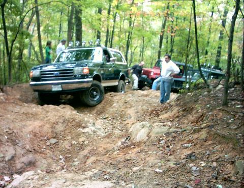 1990 Ford Bronco - A 1990 Ford Bronco tackles a challenging off-road trail in a wooded area, with other vehicles following.