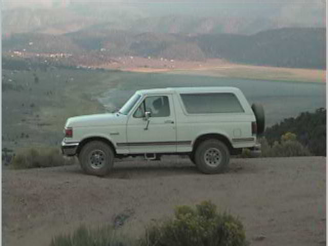 White Ford Bronco SUV side view parked on dirt overlook above a valley with mountains