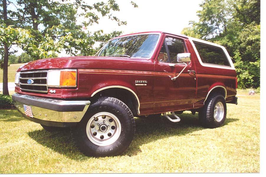 Maroon Ford Bronco SUV, front 3/4 view, parked on grass with trees behind.