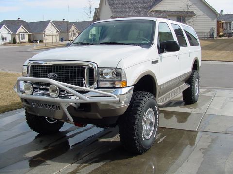 2002 Ford Excursion - This white Ford Excursion SUV features an aftermarket bumper and large tires, parked on wet pavement in front of houses.