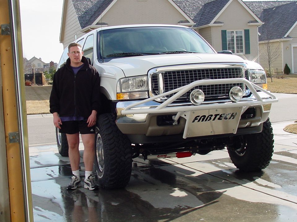 White lifted Ford Excursion with tubular bumper and man standing beside it in driveway