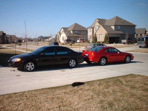 2000 Ford Taurus - A black sedan is parked next to a red coupe on a sunny day in a residential area.