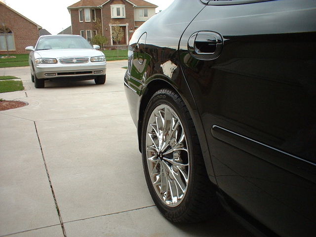 Black sedan rear quarter with chrome wheel, silver car in driveway beyond