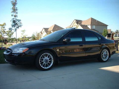 2000 Ford Taurus - This black sedan is parked in front of a suburban house. It has custom wheels and tinted windows.