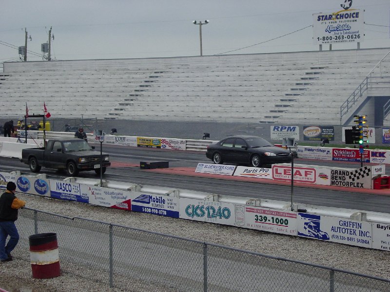 Truck and sedan on a drag strip, grandstands in background