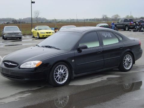 2000 Ford Taurus - A black 2000 Ford Taurus sedan is parked on wet pavement with other vehicles in the background.