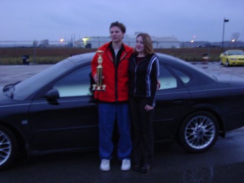 2000 Ford Taurus - Two people stand next to a black sedan, holding a trophy, with a yellow car visible in the background.