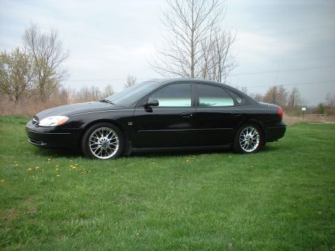 2000 Ford Taurus - A black sedan is parked on a grassy field with trees in the background under an overcast sky.