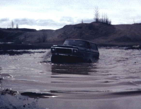Dark 1970s Ford Bronco driving through deep muddy water up to its doors