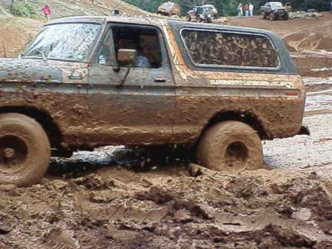 1978 Ford Bronco - A heavily mud-caked 1978 Ford Bronco struggles through deep mud, with other vehicles visible in the background.