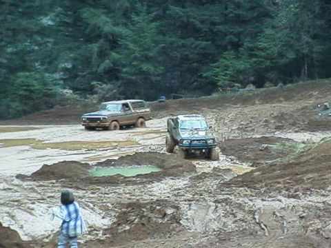 1978 Ford Bronco - A 1978 Ford Bronco and another off-road vehicle traverse a challenging muddy landscape, observed by a young child.