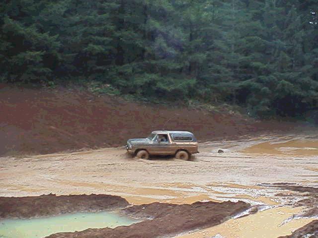 Muddy Ford Bronco crossing a clay pit beside a green water hole in the forest