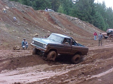 1978 Ford Bronco - Ford F-Series pickup truck driving through mud with other vehicles and people on a hill in the background.