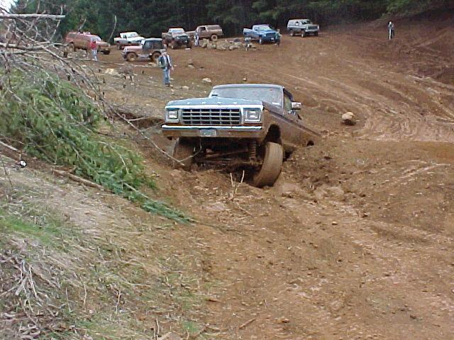 Blue 1970s Ford 4x4 pickup climbing a rutted dirt bank at an off-road park