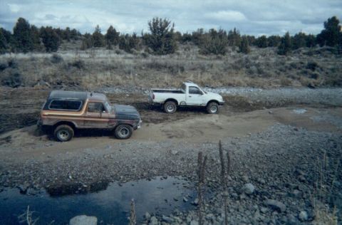 1978 Ford Bronco - Two trucks, a 1978 Ford Bronco and a white pickup, are parked on a dirt road near a body of water.