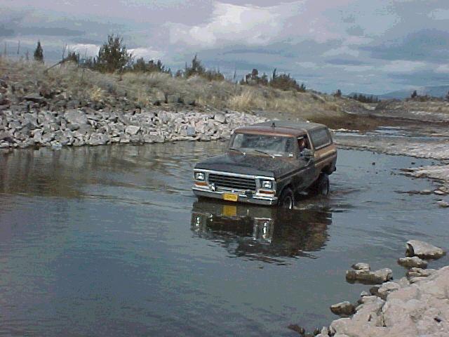 1978 Ford Bronco driving through a shallow body of water with rocky banks