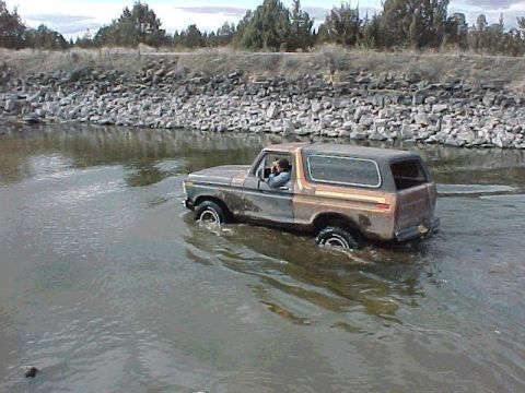 1978 Ford Bronco - A 1978 Ford Bronco navigates through a shallow body of water with a rocky embankment in the background.