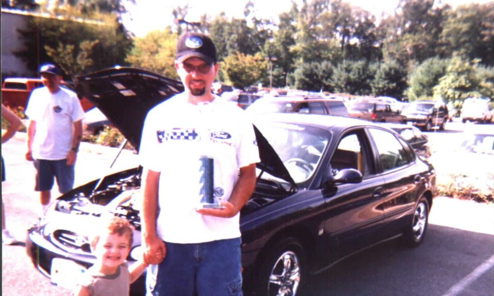 Man holding trophy and child beside dark blue sedan with open hood at a car show