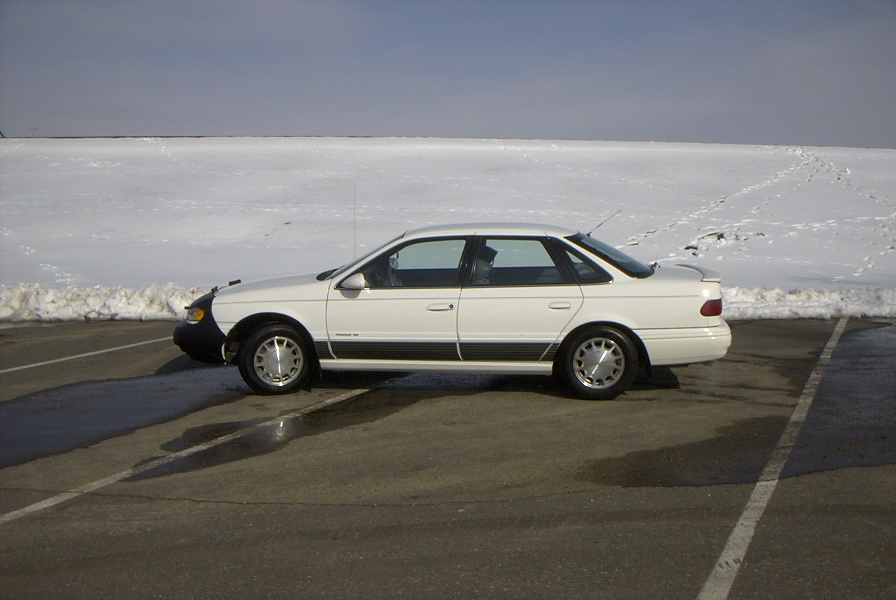 White Ford Taurus sedan with black front-end bra, side view, parked in wet lot with snowy hill behind
