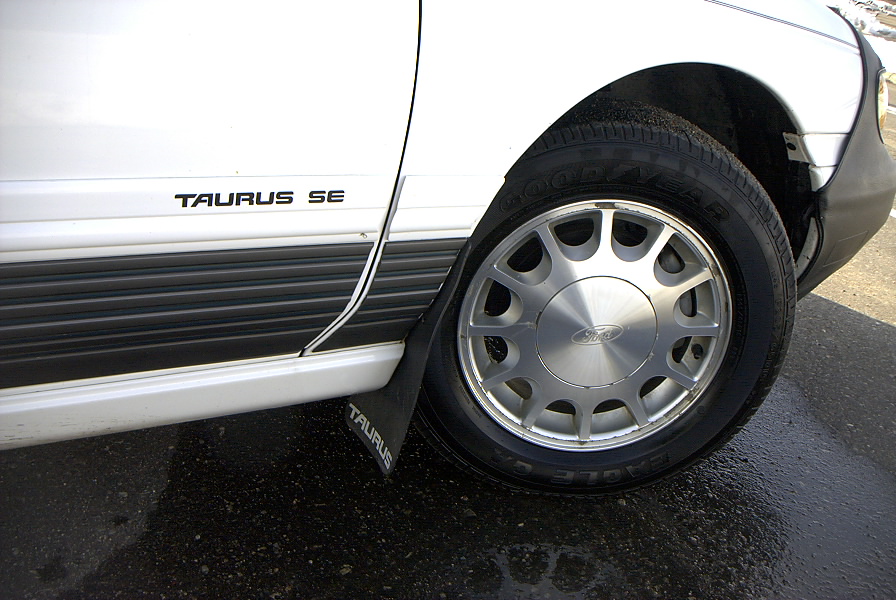 Close-up of white Ford Taurus SE front wheel with alloy rim, Goodyear tire, Taurus mud flap and SE badge