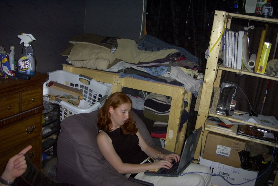 Woman with red hair using a laptop on a beanbag in a cluttered bedroom with a loft bed