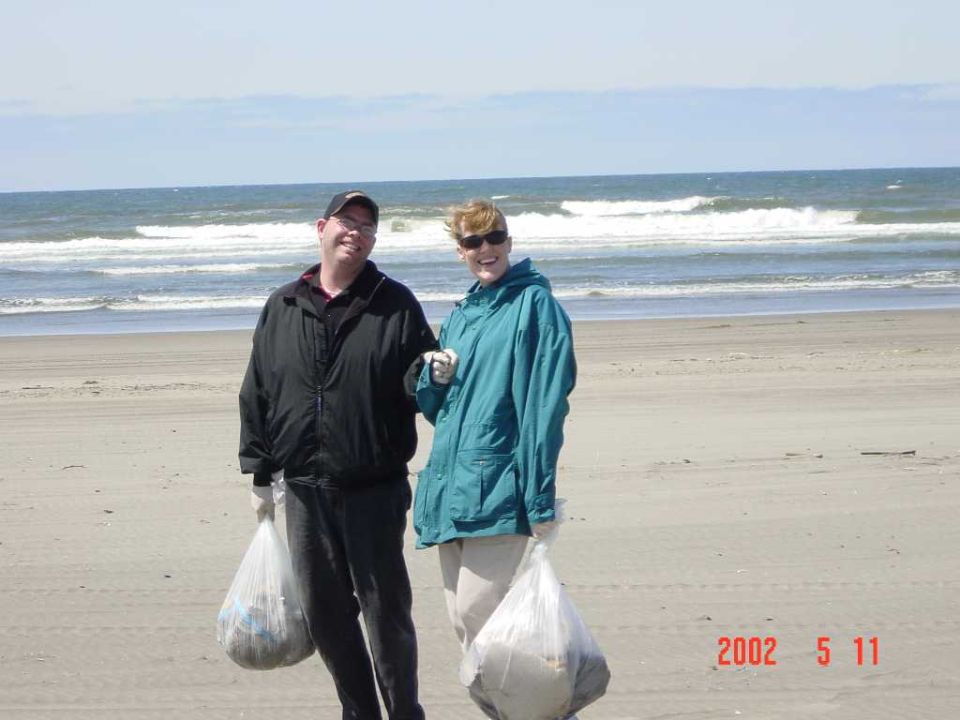 Two people holding trash bags on a beach with waves behind them, dated 2002 5 11.