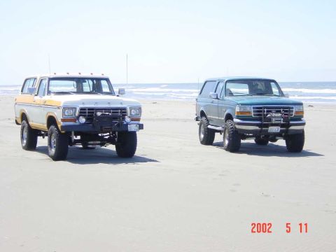 1996 Ford Bronco - Two Ford Broncos are parked on a beach, with the ocean visible behind them.