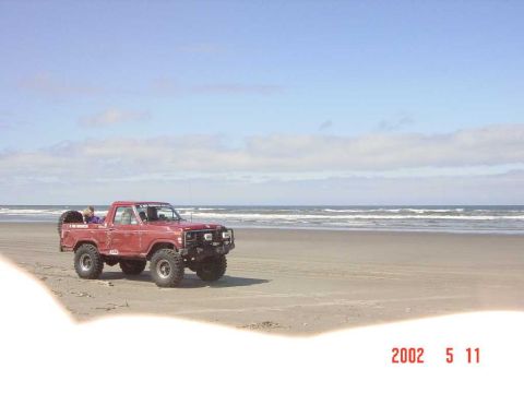 1996 Ford Bronco - A red pickup truck with off-road modifications sits on a beach with the ocean in the background.