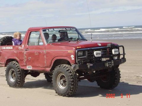 1996 Ford Bronco - A red off-road truck with large tires is parked on a beach, with text on the side indicating it belongs to the "2 BIG BRONCOS 4X4 CLUB".