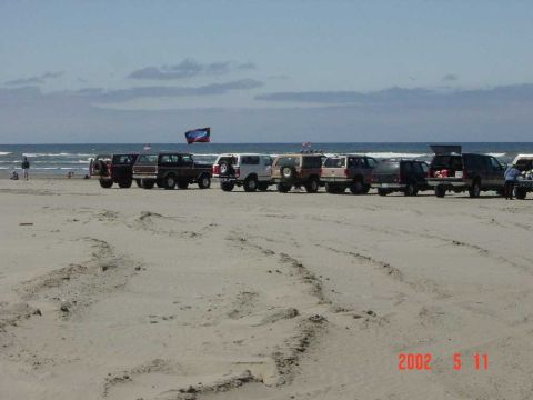 1996 Ford Bronco - A line of SUVs are parked on the sand near the ocean waves under a partly cloudy sky.
