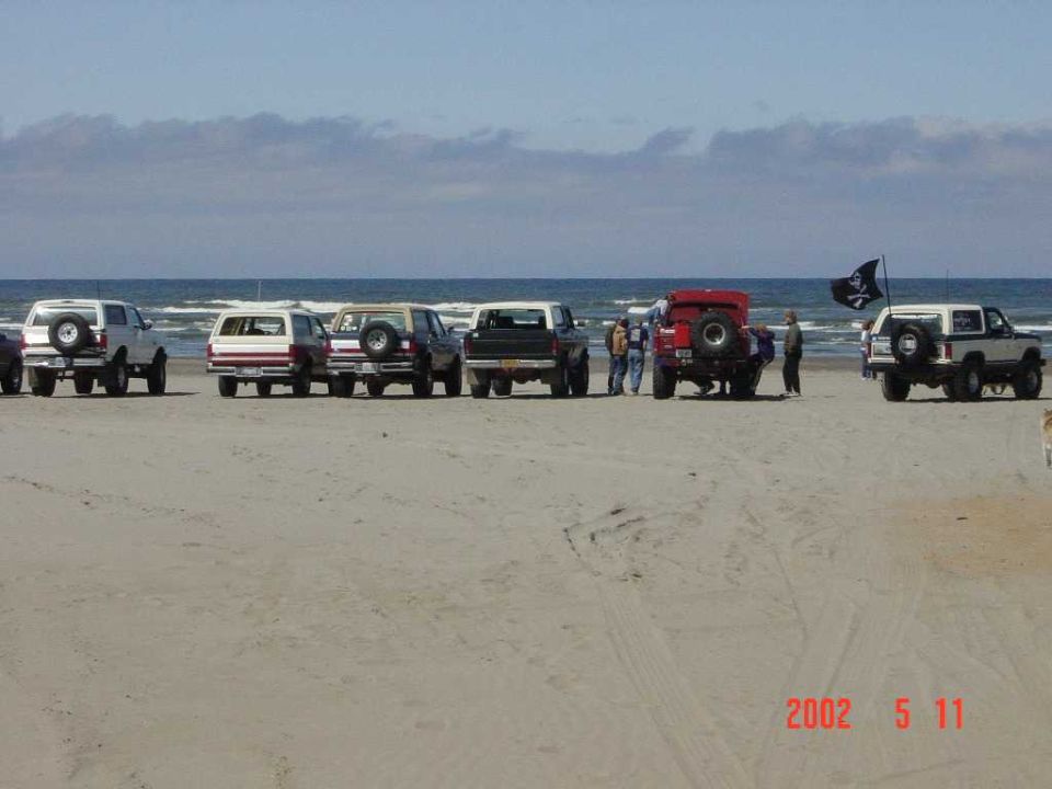 Lifted Ford Broncos and a pickup lined up on a sand beach, spare tires on rear carriers, pirate flag flying