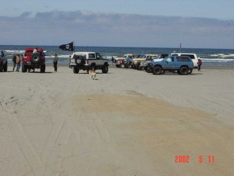 1996 Ford Bronco - A collection of SUVs and trucks are parked on a sandy beach, with the ocean in the background. A pirate flag flies from one vehicle.