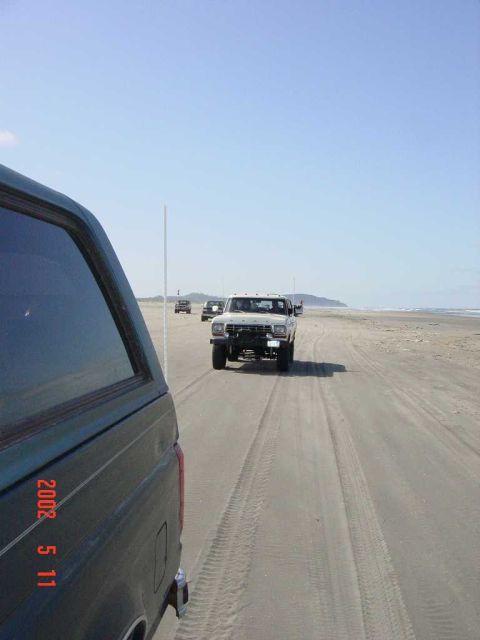 1996 Ford Bronco - Multiple Ford Broncos drive along a sandy beach under a clear blue sky.