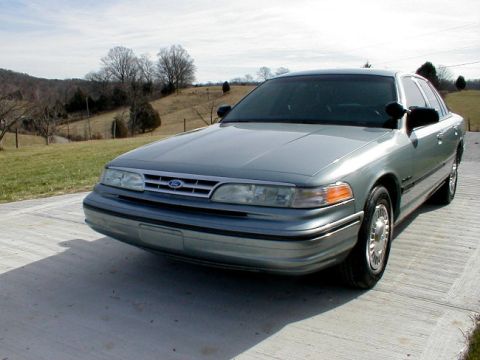 1995 Ford Crown Victoria - A silver 1995 Ford Crown Victoria is parked outside on a sunny day with a wooded hill in the background.